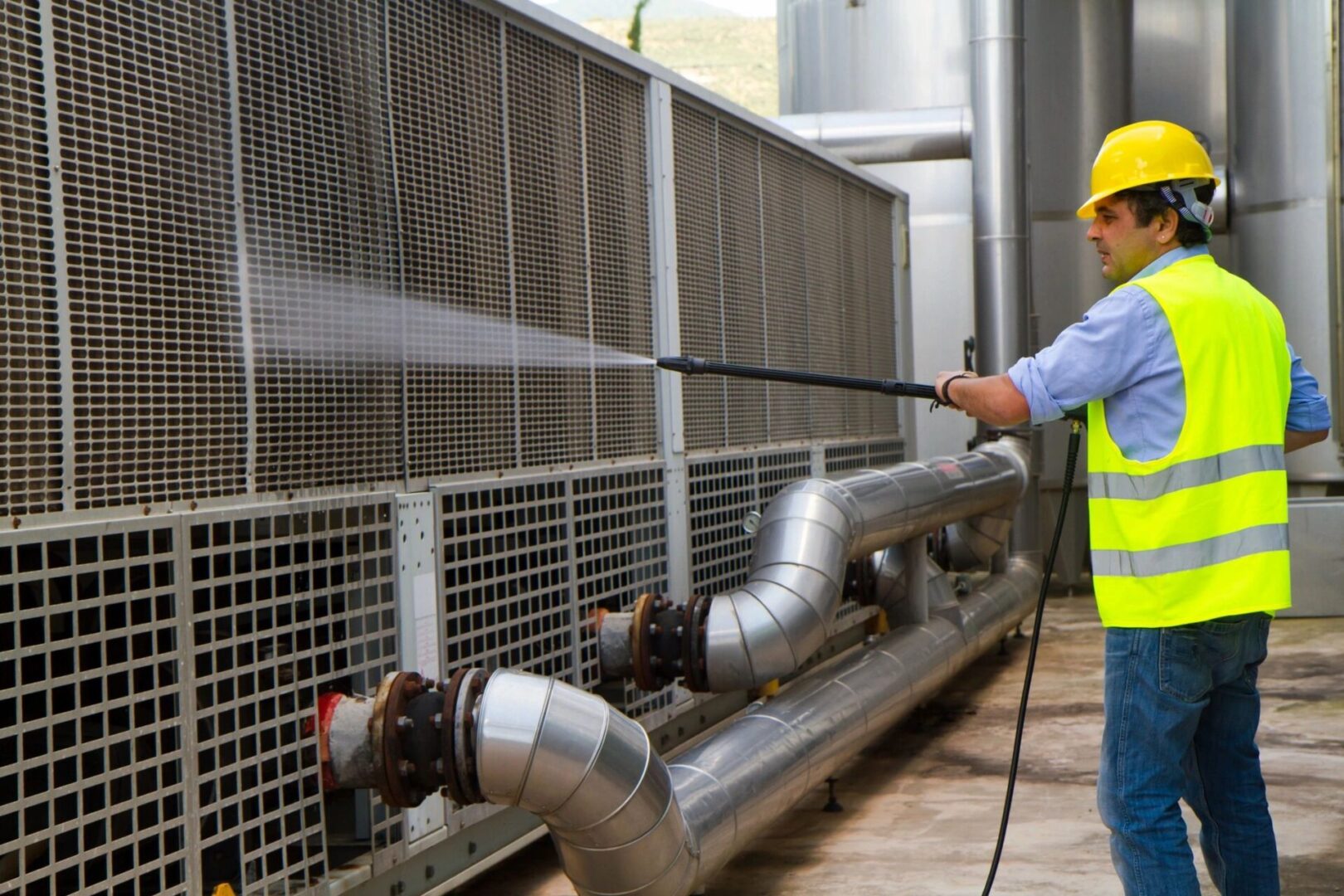 A man in yellow vest holding hose near metal pipes.