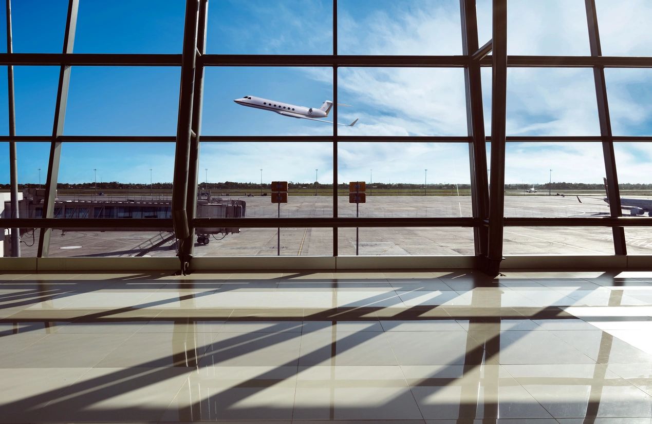 A plane is flying in the sky through an airport window.